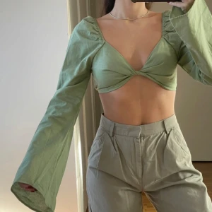 Ljusgrön croppad topp med lång ärm - Trendig ljusgrön croppad topp med twistad front och vida långa ärmar. Toppen har en djup v-ringning och smockad rygg för extra komfort. Perfekt för dig som vill sticka ut med en cool och modern stil.