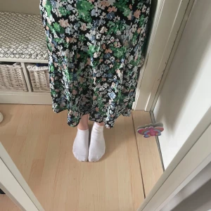 Blommig kjol från H&M - Säljer en kjol från H&M med blommigt mönster i grönt, vitt, blått och rosa på svart botten. Kjolen har en lös passform och är perfekt för varmare dagar. Superfin att styla med sneakers eller sandaler!