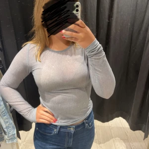 Ljusgrå glittrig topp från Kappahl - Säljer en ljusgrå glittrig långärmad topp från Kappahl.  Toppen har en tight passform och är tillverkad i ett syntetmaterial som ger en snygg glansig effekt. Perfekt för dig som vill sticka ut lite extra! Helt oanvänd med prislapp kvar! 