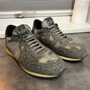 Gråa Valentino Rockrunner sneakers i ull - Säljer ett par gråa Valentino Rockrunner sneakers med kamouflagemönster. Ovandelen är i ull med detaljer i grönt skinn på hälen och grå snörning. Yttersulan är beige med nitar på baksidan. Unik och stilren design. Storlek 42! Priset går att diskutera vid snabb affär!