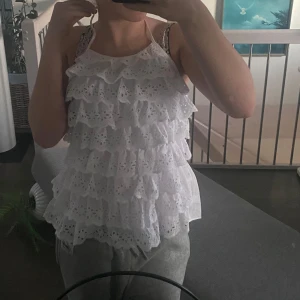 Vit volangtopp med spets - Säljer en söt vit topp med flera lager volanger och spetsdetaljer. Toppen har smala axelband och är perfekt för varma dagar. Aldrig använd 