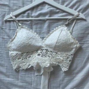 Vit spetsig croppad linnetopp - Sååå fin spets topp med snygga detaljer och gullig ”rosett” man kan knyta där bak 🤩 Använd ett par gånger men är fortfarande i bra skick! Står ingen storlek men gissar på XS/S, den är justerbar. Passar sååå bra till kjol eller jeans, perfekt till stranden! ⭐️