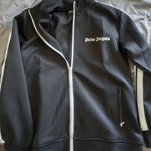 Svart track tröja från Palm Angels - Säljer en svart track tröja från Palm Angels med vit dragkedja och vita ränder längs ärmarna. Jackan har hög krage, två fickor med dragkedja och logga på bröstet. Nypris runt 4000kr säljes för 850