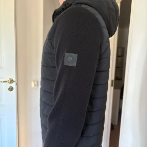 Svart cardigan från J.Lindeberg - Säljer en svart cardigan från J.Lindeberg med quiltad front och rygg samt stickade ärmar. Jackan har huva och dragkedja framtill. Perfekt för lager-på-lager och snygg till vardags.