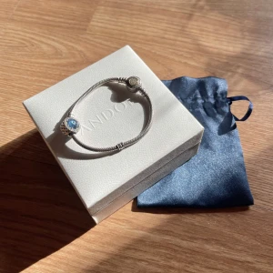 Silver armband med gulddetalj och stenar på knappen från Pandora  - Säljer ett stilrent ormkedjearmband från Pandora med en blå berlock. Silver, guld och glittrande stenar på knappen gör att det passar även fint med både silver och guld berlocker. Finns några märken men helhet och Silverfärgen är fortfarande fin, sparsamt använt. Ca17cm lång 