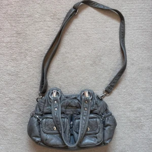 Vintage handväska - Säljer en grå axelväska från Gabarana med flera fickor framtill och bak, samt snygga silvriga detaljer. Väskan har både axelrem och handtag, och är tillverkad i syntetmaterial med quiltad look. Perfekt för dig som vill ha plats för allt viktigt.😇😇☀️pris kan diskuteras