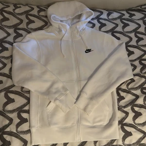 Vit hoodie med dragkedja från Nike - Säljer en klassisk vit hoodie från Nike med dragkedja och huva. Tröjan har fickor framtill och en broderad Nike-logga på bröstet. Perfekt för en sportig och avslappnad stil.