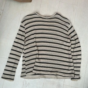 Randig stickad tröja i beige och svart - Säljer en stickad tröja med beige bas och svarta horisontella ränder. Tröjan har lång ärm och rund halsringning. 