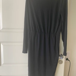 Svart långärmad klänning med resår i midjan - En stilren svart klänning med långa ärmar och resår i midjan som ger en smickrande passform. Klänningen har en rak siluett och är tillverkad i ett mjukt material, perfekt för både vardag och mer uppklädda tillfällen.