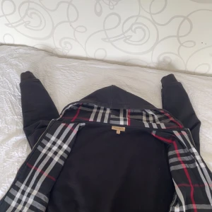 Svart hoodiejacka från Burberry - Svart hoodiejacka från Burberry med klassiskt rutigt innerfoder i svart, vitt och rött. Jackan har dragkedja framtill och broderad logga på bröstet. Perfekt för dig som gillar stilrena och tidlösa plagg med en lyxig touch.