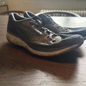 Svarta löparskor från Saucony - Svarta löparskor från Saucony med vit sula och reflekterande detalj på hälen. Skorna har snörning och är tillverkade i syntetmaterial med mesh för bra ventilation. Perfekta för löpning och träning.
