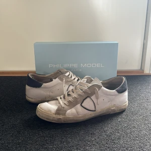 Vita och grå Philippe Model sneakers - Säljer ett par Philippe Model sneakers i vitt och grått med svarta detaljer. Skorna har snörning, rund tå och platt sula. Klassisk design med märkets logga på sidan. Perfekta för en stilren vardagslook. Nypris ca 3800, mitt pris 1200. Pris går att diskuteras om det är rimligt. Hör av er vid minsta lilla fundering🤗