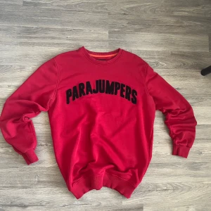 Röd sweatshirt från Parajumpers - Säljer en röd sweatshirt från Parajumpers med svart broderad logga på bröstet och patch på ärmen. Tröjan har rund halsringning och ribbade muddar vid ärmslut och nederkant. Perfekt för dig som gillar streetwear och vill sticka ut.