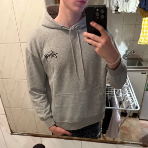 Grå hoodie från Stüssy med tärningsmotiv - Säljer en grå hoodie från Stüssy med svart logga på bröstet och stort tryck med tärningar och logga på ryggen. Tröjan har huva med dragsko och en stor magficka. Perfekt för dig som gillar streetwear.