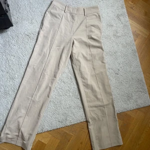 Beige kostymbyxor - Snygga beige kostymbyxor med raka ben och pressveck framtill, samt slits baktill. Byxorna har hög midja och bred linning. Perfekta till våren o sommaren för en stilren och klassisk look.