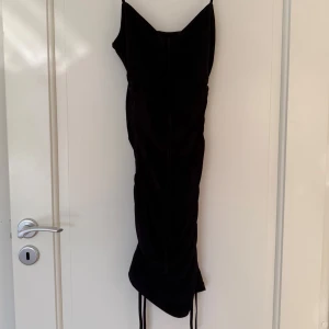 Svart tight klänning med smala axelband - Säljer en svart tight klänning med smala axelband och rynkade detaljer längs sidorna. Klänningen har en figurnära passform och är tillverkad i ett stretchigt material. Perfekt för dig som vill ha en enkel men snygg look. Tror den är från na-kd inte säker.