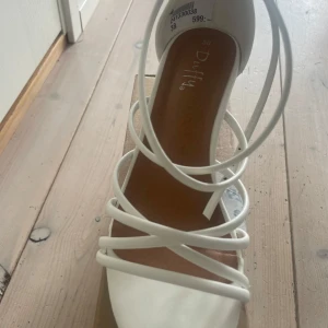 Stilrena vita skor med bred klack - Stilrena vita skor med klack från Duffy. Tunna remmar öppen tå och spänne vid ankeln. Perfekta för sommarens alla tillfällen och ger en stilren look till din outfit.