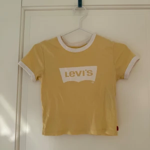 Gul t-shirt från Levi's med vit logga - Säljer en söt gul t-shirt från Levi's med vit logga på bröstet och vita kanter vid ärmar och hals. Perfekt till sommaren och enkel att matcha med jeans eller shorts.