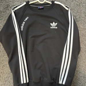 Svart sweatshirt från Adidas Neo - Svart sweatshirt från Adidas Neo med vita ränder längs ärmarna och logga på bröstet. Tröjan har rund halsringning och ribbade muddar vid ärmslut och nederkant. Klassisk och sportig stil.