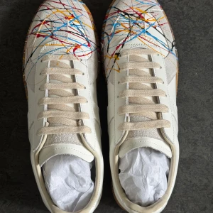 Maison Margiela sneakers med färgstänk - Unika vita sneakers från Maison Margiela med färgstänk i blått, rött, gult och svart. Skorna har beige sula, snörning och rund tå. Tillverkade i skinn och mocka för en lyxig känsla. Perfekta för dig som vill sticka ut med din stil.