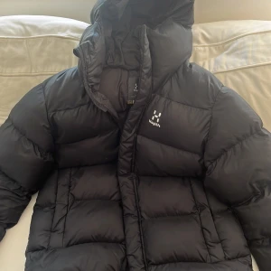 Svart dunjacka från Haglöfs - Säljer en svart dunjacka från Haglöfs i storlek L. Jackan har huva, quiltad design och dragkedja framtill. Perfekt för kalla dagar och håller dig varm hela vintern. Klassisk och stilren modell med diskret logga på bröstet. Finns litet fläck på armen men den är knappast synlig. 