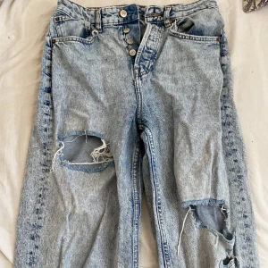 Blå jeans med slitningar - Säljer ett par blå jeans med coola slitningar på framsidan. De har en knappgylf och klassisk femficksdesign. Perfekta för en avslappnad stil.