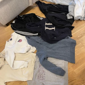 Samling av tröjor och hoodies - En samling av olika tröjor och hoodies från märken som GANT och Nike. Färgerna inkluderar svart, grå, beige och blå. Några av tröjorna har dragkedja och huva, medan andra är mer klassiska i designen. 200KR STYCK