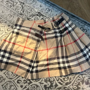  Burberry kjol - Snygg kjol från Burberry i beige, svart och röd. Kjolen har fina detljer med spännen i midjan. Säljer pågrund av ingen användning. Finns en fläck på den men som säkert kan tvättas bort🌺🌺den är för 12y men skulle kunna säga att den passar som xs också.
