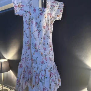 Vit blommig klänning - Charmig vit klänning med blommigt mönster i rosa och grönt. Klänningen har korta ärmar och en volangdetalj vid fållen. Perfekt för somriga dagar! Helt ny aldrig använd