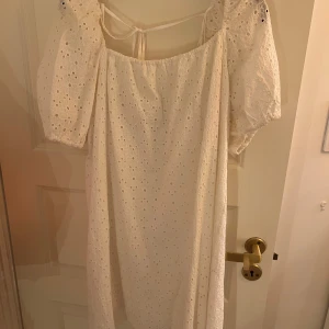 Vit klänning med puffärmar - Charmig vit klänning med puffiga kortärmar och broderade detaljer. Klänningen har en knytning i ryggen och är perfekt för sommarens festligheter.