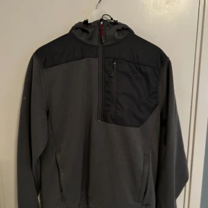 Grå övertäcks jacka från The North Face - Säljer en grå övertäcks jacka från The North Face med svarta detaljer och dragkedja. Tröjan har en praktisk bröstficka med en halv zip. Perfekt till eller från träningar, även under träningar. Funkar även att ha som jacka.