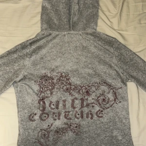 Grå hoodie från Juicy Couture - Säljer en grå hoodie från Juicy Couture med dragkedja och huva. Den har ett dekorativt mönster med blommor på. Det står M men den sitter verkligen som en Xs och kanske S men jag har s och den är tajt på mig, som man ser finns de defekter vid ärmarna då de är slitna där, därav priset, jätte fin annars, vinröda diamanter på ryggen som alla finns kvar och trycket är synligt, möjligtvis lite utsuddat men man ser hur de är på bilden💕en gammal juicy med den gamla modellens J på dragkedjan, missfärg ❌