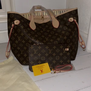 Brun neverfull väska - Säljer en snygg brun axelväska med det klassiska monogrammönstret. Väskan har ljusa handtag och detaljer i beige med röda kanter. Insidan är randig med en praktisk dragkedjeficka. Perfekt för att bära med sig det viktigaste med stil.