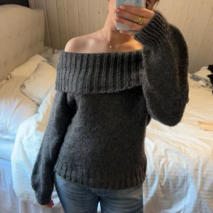 Mörkgrå stickad offshoulder-tröja - Säljer en mysig mörkgrå stickad tröja med offshoulder-design. Tröjan har långa ärmar och en bred ribbad kant vid axlarna som ger en stilren look. Perfekt för kyliga dagar.