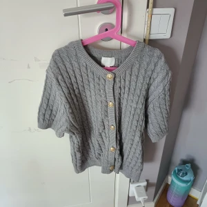 Grå stickad kofta från H&M - Säljer en snygg grå stickad kofta från H&M med kabelstickat mönster och guldknappar framtill. Perfekt för kyliga dagar och ger en stilren look. Passar bra till både jeans och kjol. Den är i storlek L men skulle säga att den passar folk mellan S och L! Då jag har S igentligen 