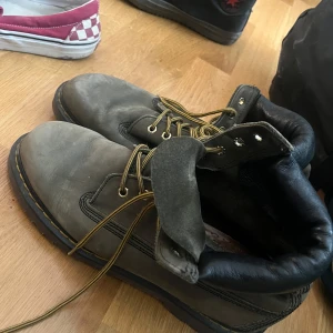 Grå boots med snörning från Dr. Martens - Säljer ett par robusta grå boots med snörning och svarta detaljer. Perfekta för en tuff och stilren look. De har en rund tå och är gjorda för att hålla länge.