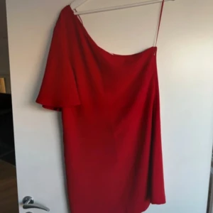 Röd one-shoulder klänning - Snygg röd one-shoulder klänning perfekt för festliga tillfällen. Klänningen har en elegant asymmetrisk design med en kort ärm på ena sidan. Den är tillverkad i ett mjukt och bekvämt material som faller fint.