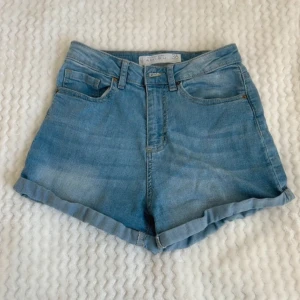 Blå jeansshorts - Säljer ett par klassiska blå jeansshorts med upprullade kanter. De har en bekväm passform och är perfekta för sommardagar. Shortsen har fem fickor och en knappgylf.
