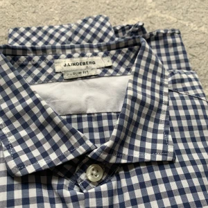 Blå J.Lindberg skjorta  - Snygg blå och vit rutig skjorta från J.Lindeberg i slim fit. Skjortan har långa ärmar och knappar framtill. Perfekt för en stilren look med en liten broderad logga på bröstet.