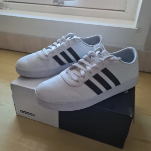 Vita Adidas Easy Vulc 2.0 sneakers - Säljer ett par vita Adidas Easy Vulc 2.0 sneakers med klassiska svarta adidas ränder. Skorna har snörning och en stilren design som passar perfekt till vardags. De är bekväma och lätta, vilket gör dem idealiska för en snygg look. Sparsamt använda. Storlek 43 1/3. Kan hämtas i Vallentuna.