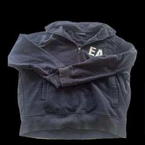 Svart hoodie från Emporio Armani - Säljer en stilren svart hoodie från Emporio Armani med dragkedja och EA-logga på bröstet. Perfekt för en avslappnad look. Tröjan har långa ärmar och en bekväm passform.