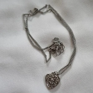 Silverfärgat halsband med hjärthänge - Elegant silverfärgat halsband med ett vackert hjärthänge. Hänget har ett intrikat mönster som ger en romantisk känsla. Perfekt som en subtil accessoar för att komplettera din look.