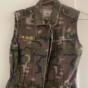 Camouflageväst  - Snygg camouflageväst från Brave Soul med coola stjärndetaljer och nitar på kragen. Västen har knappar framtill och praktiska fickor. Det är storlek 36/XS