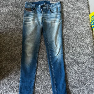 Blå lågmidjade jeans från Diesel - Snygga blå lågmidjade jeans från Diesel med en klassisk femficksdesign och en lätt tvättad look. De har en knappgylf och en liten Diesel Industry-detalj på fickan. Perfekta för en avslappnad stil. Storlek W 25 L 32