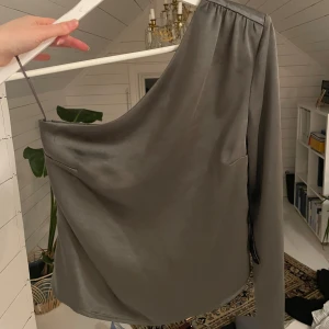 Grå one shoulder topp från Gina Tricot - Snygg, grå one shoulder topp i satin från Gina Tricot. Helt oanvänd. Säljs pga aldrig kommit till användning. Skimmrar superfint i verkligheten.