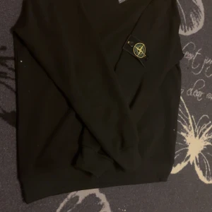 Svart tröja från Stone Island - Säljer en stilren svart tröja från Stone Island med deras ikoniska märke på ärmen. Tröjan har långa ärmar och en klassisk passform. Perfekt för en avslappnad och trendig look.