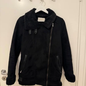 Svart jacka från Vila - Säljer en snygg svart jacka från Vila med en mysig fuskpälsfoder. Jackan har en asymmetrisk dragkedja och två fickor med dragkedjor. Den har en hög krage och justerbara spännen vid midjan för en perfekt passform.