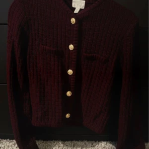 Vinröd stickad kofta från H&M - Säljer en vinröd stickad kofta från H&M med dekorativa guldknappar. Koftan har en ribbad struktur och långa ärmar. Perfekt för kyliga dagar!