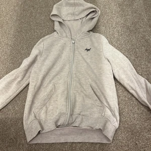 Beige hoodie med dragkedja - Säljer en stilren beige hoodie från Hype med dragkedja framtill. Tröjan har en luva och långa ärmar, perfekt för en avslappnad look. Den har ribbade muddar vid ärmslut och nederkant.