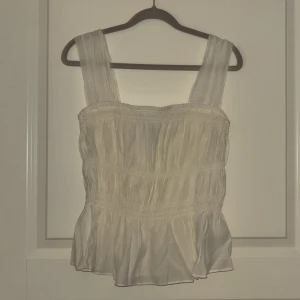 Vit blus/linne från H&M - Säljer en söt vit blus/linne från H&M med breda axelband och smockdetaljer. Perfekt för sommaren. Blusen har en luftig passform och är tillverkad i ett lätt material. Pris kan diskuteras 